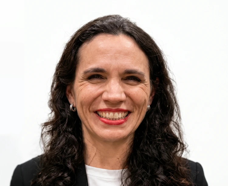 Portrait of a woman smiling with long dark curly hair in a black blazer and white shirt on a plain background.