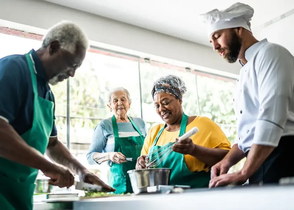 Seniors and staff members wearing aprons prepare food together in a bright senior living community kitchen.