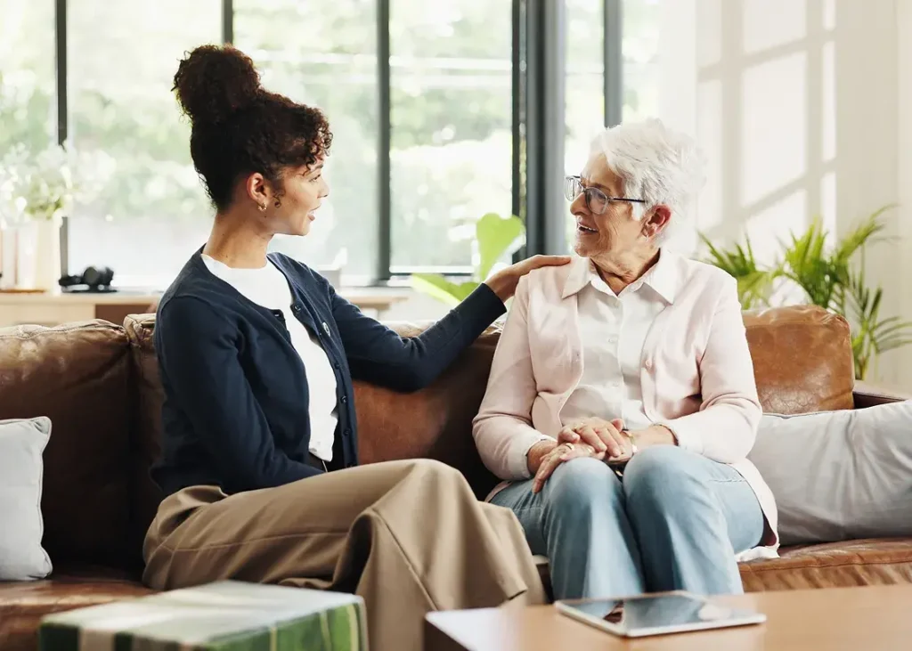 Two women sitting on a couch in a bright living room sharing a warm conversation with one woman gently touching the other's shoulder.