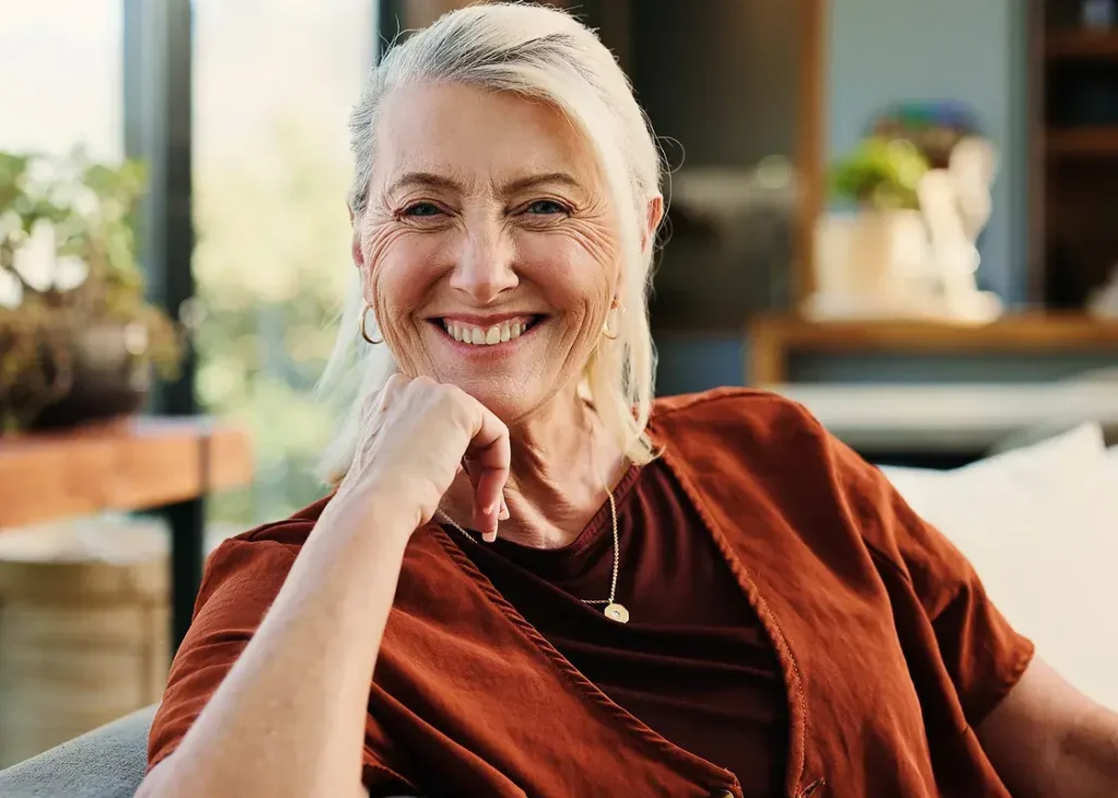 Smiling older adult woman sitting in a cozy indoor living space with natural light and plants, representing a warm community lifestyle.
