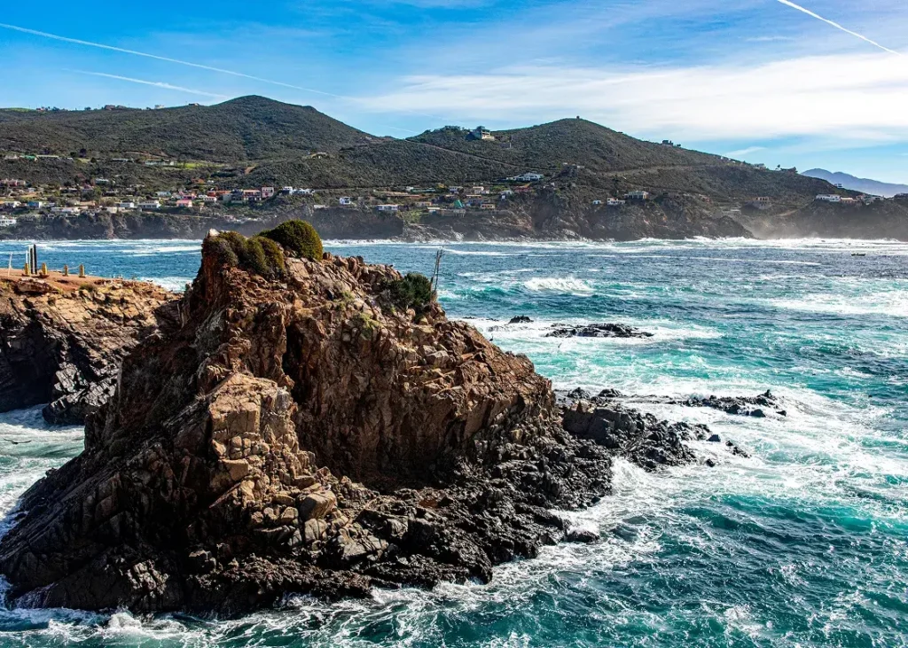 Rocky coastline with ocean waves crashing and hillside houses in the distance under a clear blue sky.