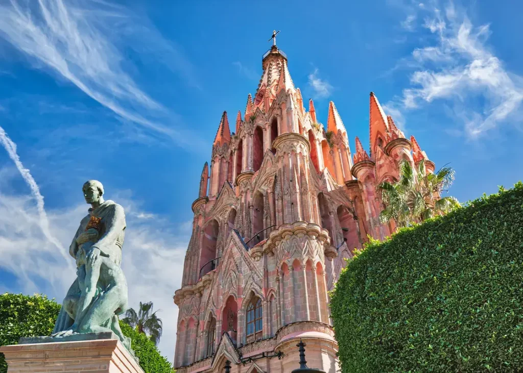 Ornate historic building with spires and arched windows beside a bronze statue, set against a bright sky with clouds and surrounding greenery.