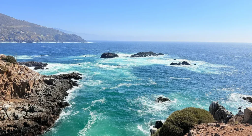 Rocky coastal cliffs overlooking waves crashing into the ocean under a clear blue sky.