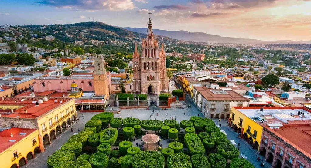 Aerial view of a historic plaza with a gothic cathedral and manicured greenery surrounded by colorful buildings at sunset.