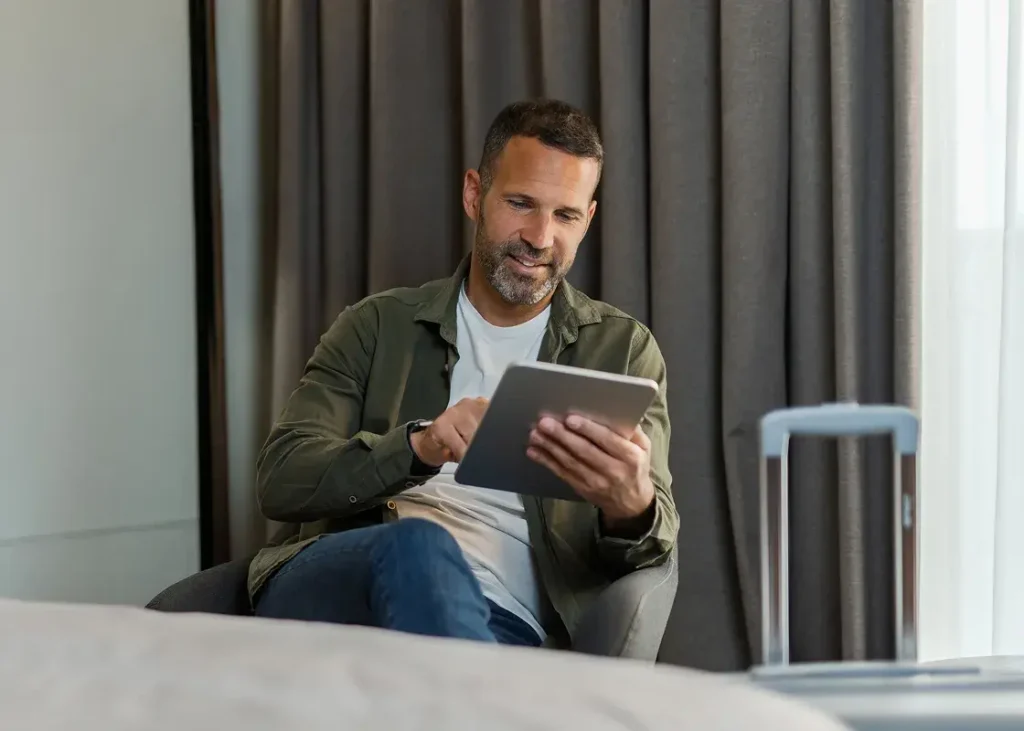 Man sitting indoors in a chair using a tablet with a suitcase nearby, next to curtains.