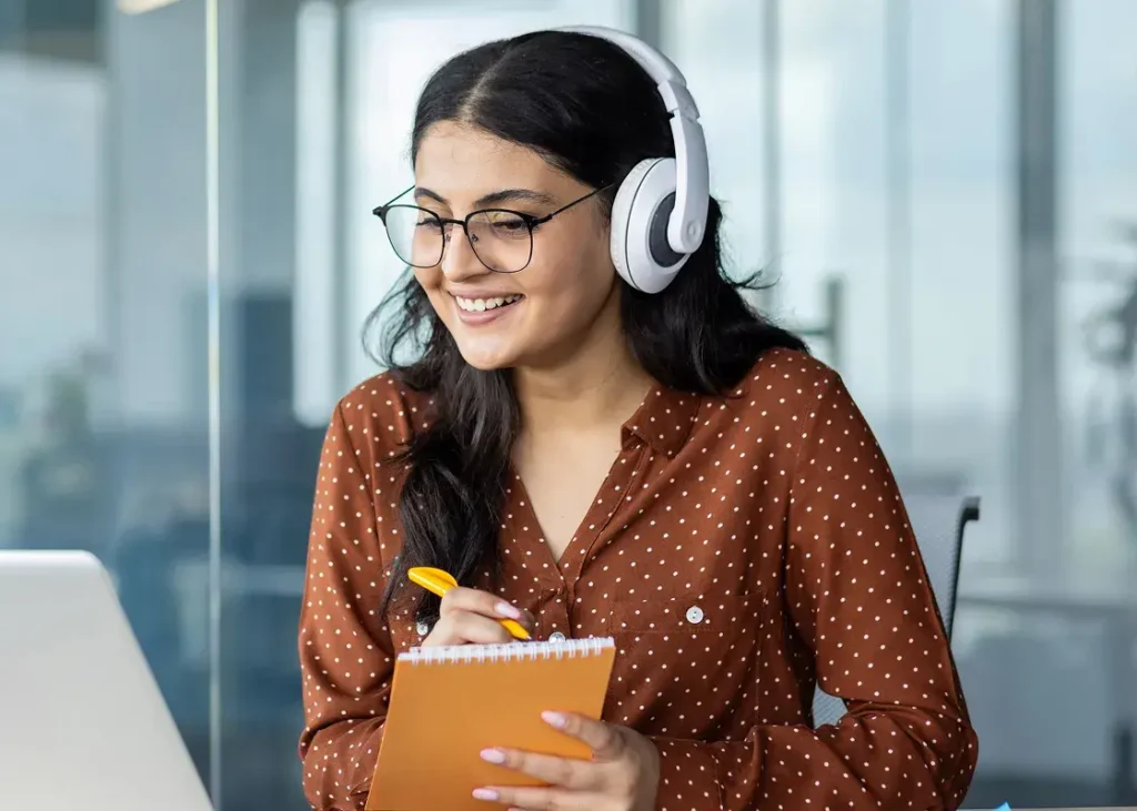 Smiling woman wearing glasses and headphones engaged in an online meeting while taking notes in a modern office setting.