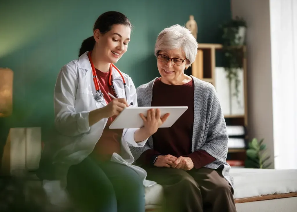 A healthcare professional shares information on a tablet with an elderly woman seated indoors in a bright room with plants and shelves.