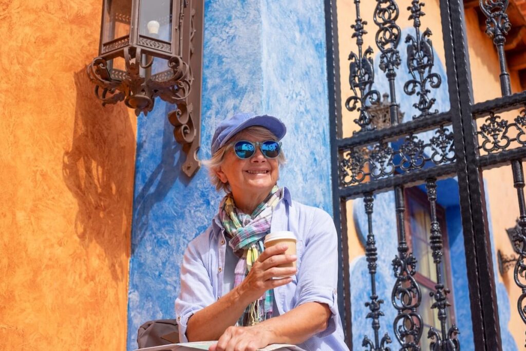 A woman sitting outdoors near a colorful building, smiling while holding a coffee cup, with decorative ironwork and a wall lantern visible.