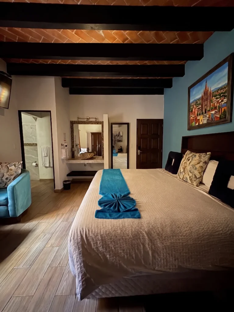 Comfortable bedroom with a large bed, decorative pillows, blue bed runner, armchair, and framed artwork under exposed beam ceiling in a senior living community.