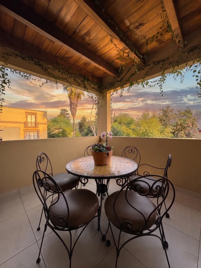 Covered outdoor patio with a round mosaic table, iron chairs with cushions, and a potted plant under a wooden ceiling with vines, overlooking trees and a sunset sky.