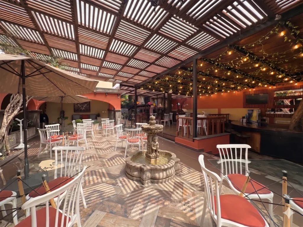 Outdoor dining area with white chairs and tables under a pergola with string lights and a central decorative water fountain enhancing the community atmosphere.