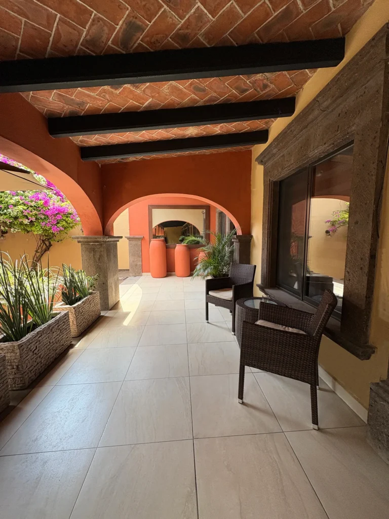 Covered outdoor seating area with wicker chairs, glass-top table, and potted plants under a terracotta tile roof with wooden beams.