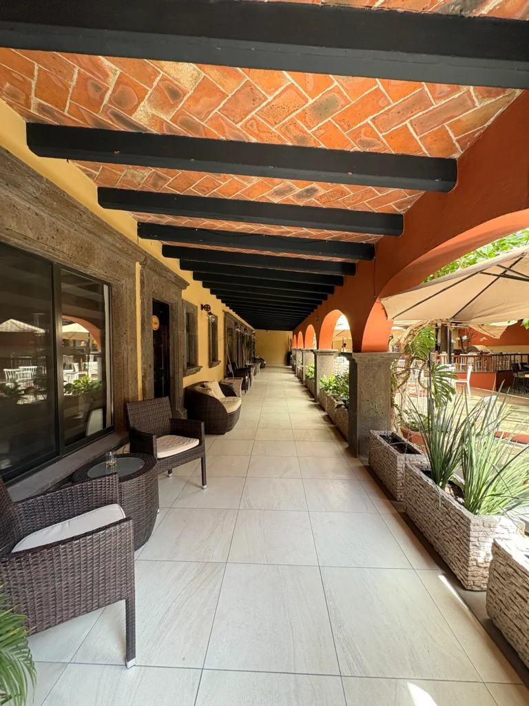 Covered outdoor corridor with seating and plants overlooking a courtyard with tables and umbrellas in the community.