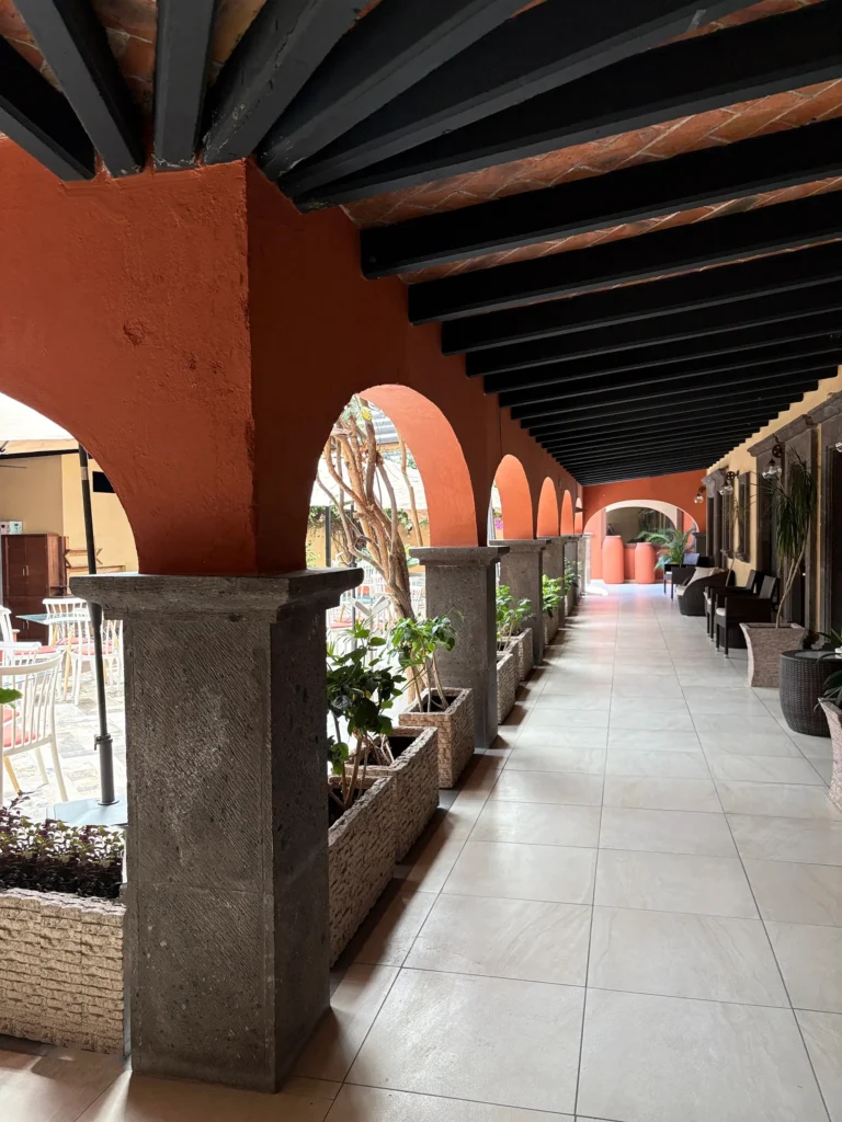 Covered outdoor corridor with arched walls, stone pillars, plants, and seating areas creating a peaceful amenity space.
