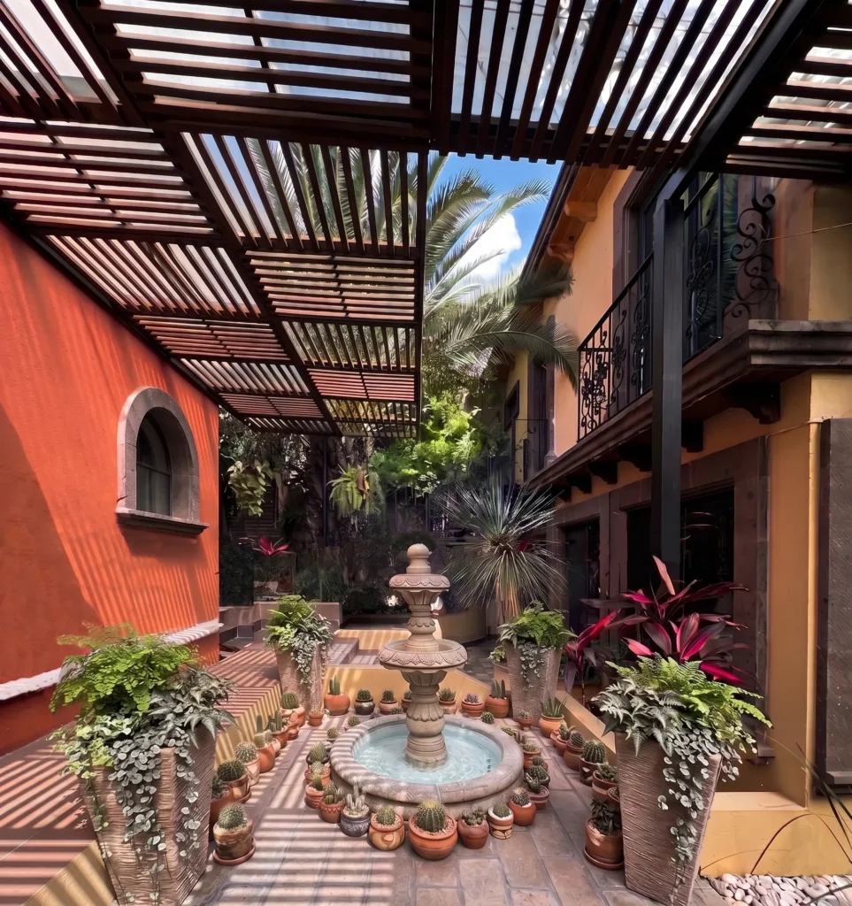 A shaded courtyard featuring a tiered water fountain surrounded by a variety of potted plants and succulents between two buildings with balconies and tropical greenery.