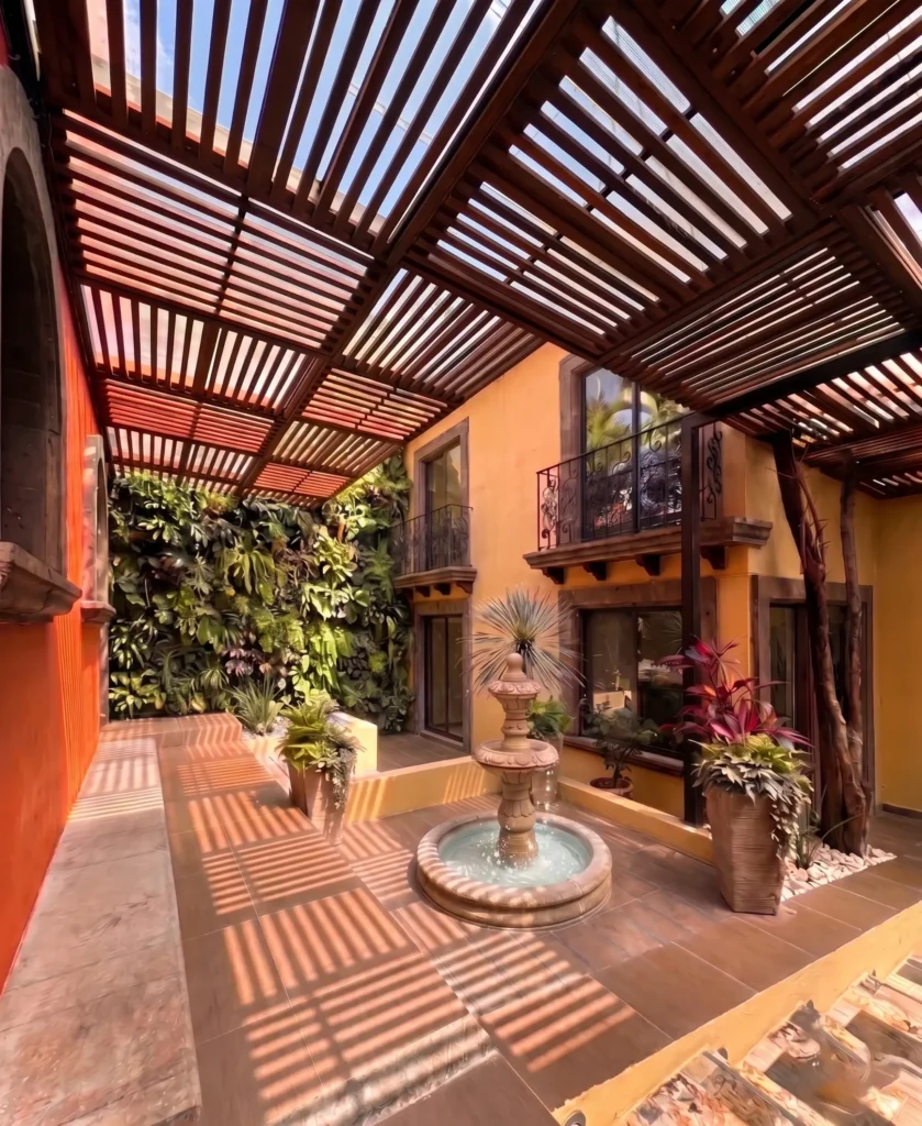 Outdoor courtyard at Serenity Bridges featuring a water fountain, lush potted plants, a vertical garden wall, and a wooden pergola casting patterned shadows on the tiled floor.
