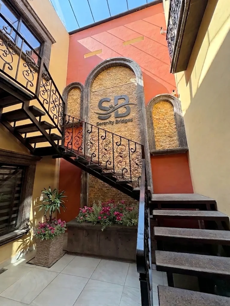Indoor stairwell with decorative iron railings, planter with pink flowers, and Serenity Bridges logo on a textured wall.