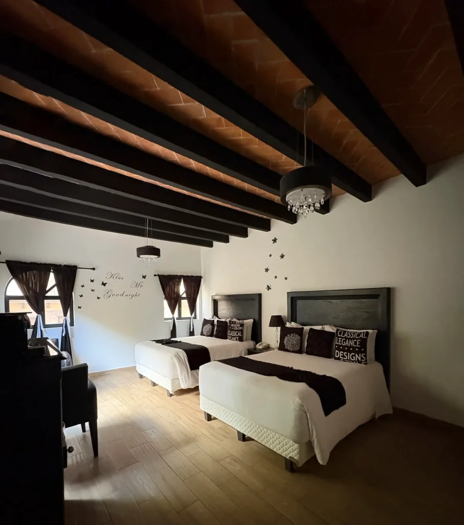Bedroom with two beds, wooden beam ceiling, white bedding with dark accents, and natural light from windows with dark curtains.