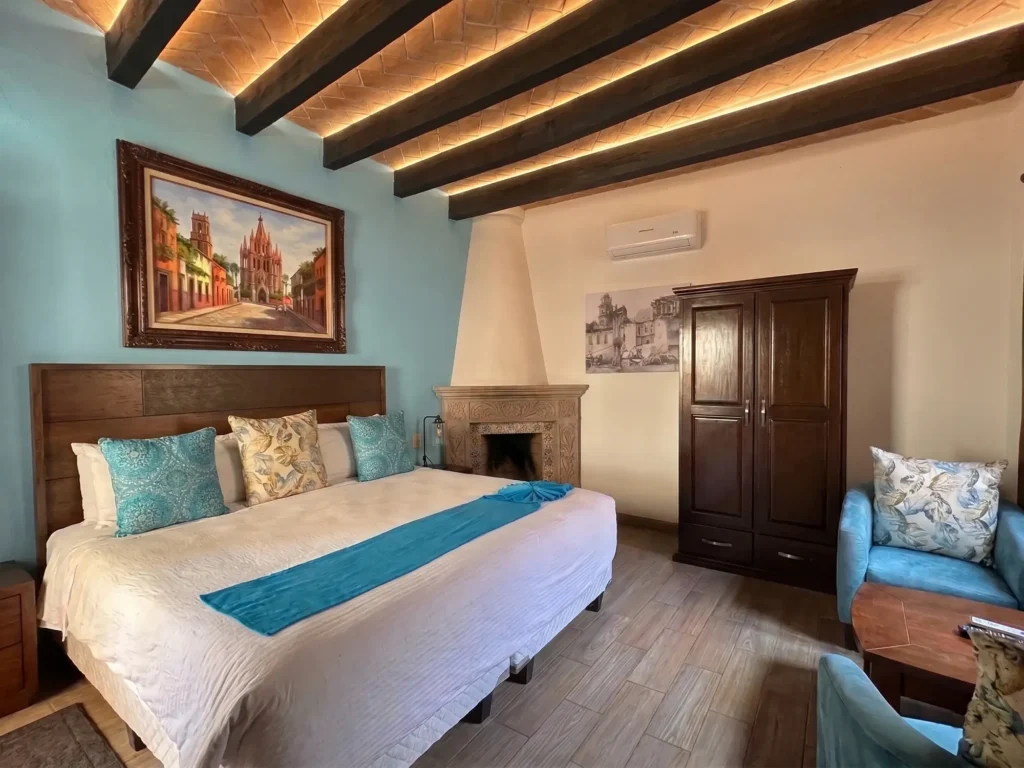 Cozy bedroom with a large bed, decorative pillows, wooden furniture, a fireplace, and framed artwork in a warm, inviting setting.