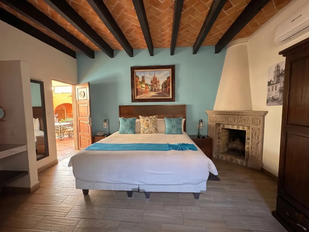 Bedroom with a neatly made bed with blue accents, a decorative fireplace, wooden ceiling beams, and an open door to an outdoor patio in a senior living community.